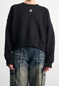 Musta lyhythihainen collegepaita, jossa on pyöreä kaula-aukko ja pitkät hihat; edessä pieni valkoinen Adidas-logo brodeerattuna rintaan. Yhdistettyinä sinisiin farkkutyyppisiin cargo-housuihin.