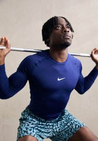 Marineblaue Sportlongsleeve mit weißem Logo; gemusterte grüne Shorts; individuelle Kniebeuge mit einer Langhantel auf den Schultern.