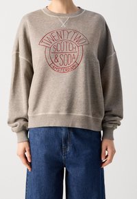 Sudadera gris con cuello redondo y mangas largas, con un logotipo circular rojo que dice "TWENTY TWO SCOTCH & SODA AMSTERDAM".