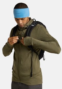 Olivgrüner Zip-Hoodie aus leichtem Stoff, mit Daumenlöchern in den Ärmeln, getragen mit einem schwarzen Rucksack und einem hellblauen Haarband.