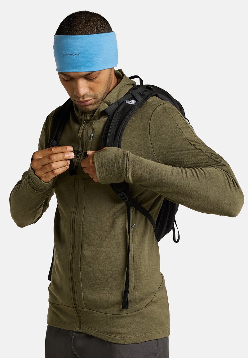 Olivgrüner Zip-Hoodie aus leichtem Stoff, mit Daumenlöchern in den Ärmeln, getragen mit einem schwarzen Rucksack und einem hellblauen Haarband.