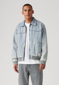 Veste en denim bleu clair avec fermeture éclair à l'avant, comportant deux poches, des poignets côtelés et une texture légèrement délavée. Portée par-dessus un t-shirt blanc.