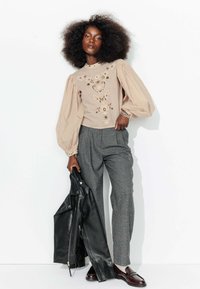 Blouse beige brodée avec des manches bouffantes ; pantalons à carreaux gris ; tenant une veste en cuir noire ; mocassins marron ; fond blanc.