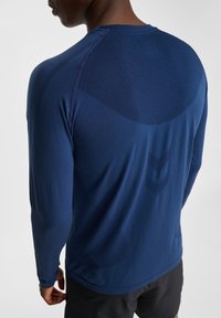 Mörkblå långärmad atletisk topp med en slät yta, som har meshpaneler på baksidan och subtil märkning i form av en chevron.