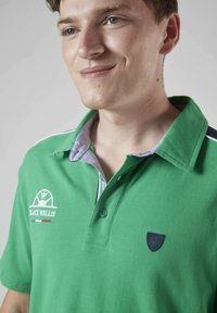 Polo vert en tissu texturé, avec un col bleu et un logo brodé. Les accents notables incluent des détails blancs et rouges.