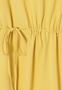 Tejido amarillo con una textura suave, que presenta lados elásticos fruncidos y detalles atados, creando un aspecto ajustado en la cintura.