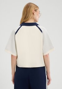 Kobieta z długimi blond włosami i okularami, ubrana w krótką bluzkę w kolorze kremowym i granatowym oraz granatowe spodnie, pokazana z tyłu.