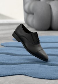 Chaussure à lacets en cuir noir avec une texture lisse, un bout arrondi et des détails subtils sur la tige, présentée sur un tapis bleu texturé.