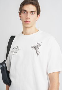 Biele bavlnene tričko s čiernym grafickým motívom krídlatej postavy a značkou Nike na ľavej hrudi; model nesie čiernu tašku.