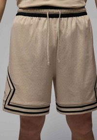 Pantalones cortos deportivos beige con textura perforada, cintura elástica negra, cordón ajustable y rayas negras en contraste a lo largo de los lados.