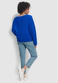 Un maglione blu oversize con una finitura testurizzata, abbinato a jeans azzurri chiari e scarpe da ginnastica bianche con dettagli dorati. Vista posteriore.