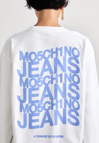 Balta garām piedurknēm krekls ar uzdruku, kurā bolds zils teksts "MO5CH1NO JEANS" atkārtojas vairākkārt, ar papildu saukli zem tā.