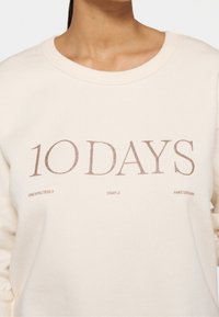 Sweat-shirt beige clair avec col rond, arborant un texte doré métallique "10 DAYS" et un texte plus petit "UNEXPECTEDLY SIMPLE AMSTERDAM." Texture douce.