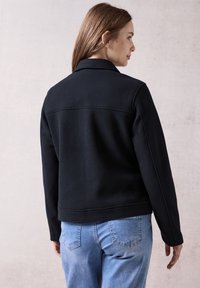 Veste en polaire noire avec un col, des coutures structurées et une coupe décontractée. Associée à un denim délavé clair, mettant en avant un look simple et informel.