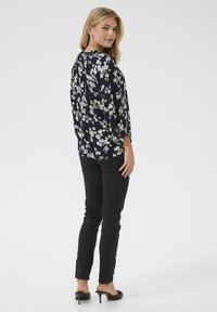 Marinebluse mit floralen Muster in Weiß, langen Ärmeln und gerafftem Ausschnitt; kombiniert mit schwarzen Skinny-Jeans und schwarzen Absatz-Mules.