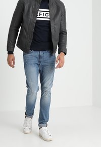 Černá kožená bunda s prošívanými ramenními částmi, námořnická mikina s bílým textem, světle modré prodřené džíny a bílé tenisky.