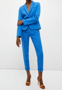 Blå skräddarsydd kostym med en figursydd kavaj och smala byxor, tillverkad av slätt material. Har uppvikta ärmar och orangea remsandaler.