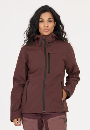 Vrouw met lang donker haar draagt een bordeauxrode jas met capuchon en bijpassende broek, staat met één hand in haar zak tegen een effen achtergrond.