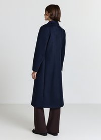 Marinblauer Wollmischmantel mit hohem Kragen, geradem Schnitt und langen Ärmeln. Glatte Textur und minimalistisches Design, kombiniert mit braunen Hosen.