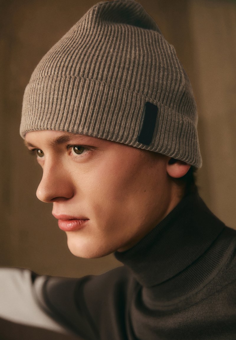 Gorro cinza canelado com a bainha dobrada e uma pequena etiqueta preta. Usado por um modelo com um suéter de gola alta, destacando a sua textura e ajuste.