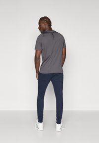 Grå kortermet t-skjorte med en vertikal svart logo på baksiden, kombinert med marineblå joggebukser, vist fra baksiden, med hvite joggesko.