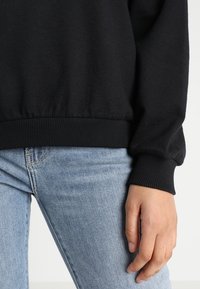 Felpa nera con polsini a costine, tessuto morbido e vestibilità rilassata, abbinata a jeans blu. Focus sulla texture e sui dettagli di design senza soluzione di continuità.
