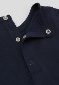 Námořnická modrá pletená košile s límečkem, která má stříbrné patentky a hladkou texturu. Detailní záběr na výstřih a detaily knoflíků.