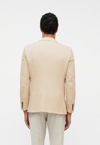 Άνδρας με σγουρά σκούρα μαλλιά που φοράει μπεζ σακάκι και ανοιχτό γκρι παντελόνι, απεικονιζόμενος από την πίσω πλευρά σε απλό λευκό φόντο.