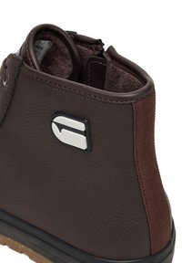 Tuva brūna ādas potītes zābaka ar polsterētu apkakli, auduma papēža paneli, melnu zoli un baltu taisnstūra formas logo sānos.