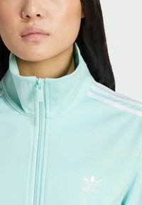 Giacca zip-up azzurro chiaro realizzata in tessuto morbido, con colletto alto e accenti bianchi a tre strisce sulle spalle, con un piccolo logo sul petto.