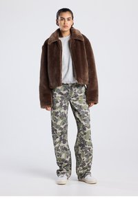 Ruskea tekoturkistakki, joka on lyhyt ja siinä on korkea kaulus, yhdistetty vihreään maastokuvioituun housuun. Malli käyttää valkoisia lenkkareita ja vaaleanharmaata toppia.