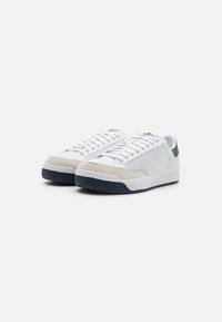 White sneakers with a textured fabric upper, grey suede accents, and a navy rubber sole. Laces are white with blue branding on the tongue.