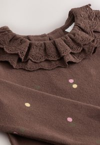 Ruskea kangasvaate, jossa on röyhelöinen pitsikaulus. Pinnalla on värikkäitä pyöreitä kuvioita vaaleanpunaisen, keltaisen ja vihreän sävyissä.