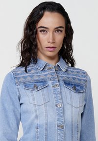 Kék farmerdzseki díszes varrott mintákkal a vállakon, két elülső zsebbel és ezüst színű patentgombokkal.