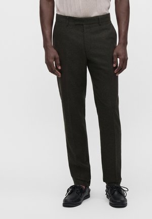 Homme portant un pantalon vert foncé sur mesure et des chaussures en cuir noires à lacets, debout devant un fond clair uni.