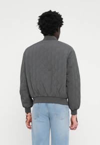 Γκρι καπιτονέ μπουφάν βόμβας με ριμπ μανσέτες και στρίφωμα. Διαθέτει έναν απλό σχεδιασμό, λεία υφή και χαλαρή εφαρμογή. Παρουσιάζεται θέα από πίσω.