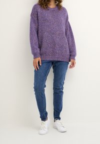 Lila stickad tröja med lös passform och ribbad manschett, kombinerad med blå jeans och vita sneakers. Strukturert tyg med flerfärgade prickar.