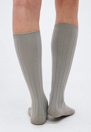 Dolne części nóg w szarych, prążkowanych podkolanówkach do kolan, stojące na jasnej powierzchni, z jedną piętą lekko uniesioną.