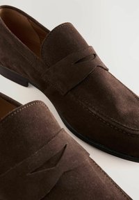 Mocassins en daim marron avec une semelle plate, présentant une découpe décorative et des coutures contrastantes le long du bord. Doublure intérieure lisse.