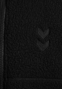 Zbliżenie na czarną tkaninę o fakturze z pionowym zamkiem błyskawicznym po lewej stronie i dwoma naszytymi kształtami w formie chevronu skierowanymi w dół po prawej stronie.