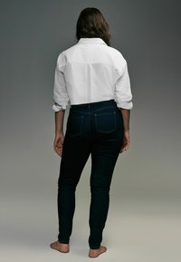 Camisa blanca de botón con mangas dobladas, combinada con jeans ajustados azul oscuro. Descalzo, visto desde atrás sobre un fondo neutro.