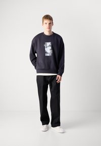 Sweatshirt blu scuro con grafica anteriore, vestibilità ampia, polsini e orlo ribattuti, abbinato a pantaloni neri e sneakers bianche.