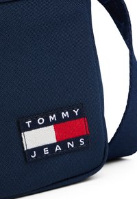 Borsa a spalla in tessuto blu navy con un patch ricamato rettangolare che riporta la scritta "TOMMY JEANS" e un logo rosso, bianco e blu.