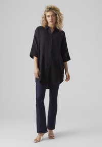 Camicia nera a bottoni con vestibilità relaxed, maniche a tre quarti e tessuto testurizzato, abbinata a jeans a zampa d'elefante blu scuro e tacchi bianchi.