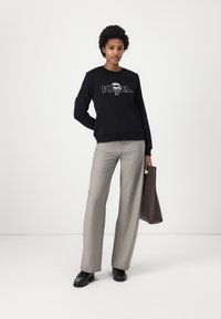 Črna kapuca s grafičnim logotipom na sprednji strani, v kombinaciji s svetlo sivo širokimi hlačami in rjavim torbicami. Usnjeni čevlji dopolnjujejo videz.