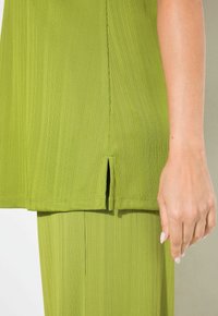 Top in tessuto verde a coste con una fessura laterale e una vestibilità rilassata. La texture è liscia, con strisce verticali, abbinato a pantaloni coordinati.