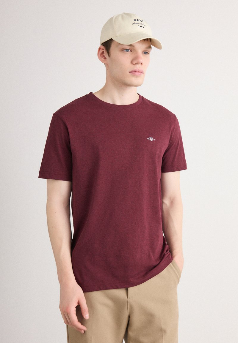 Jonge man met een beige pet, bordeauxrood t-shirt met een klein logo op de borst, en beige broek, staand met één hand in zijn zak tegen een effen achtergrond.