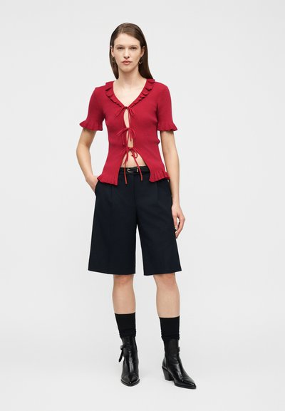Mujer con una blusa roja de manga corta con tres lazos en la parte delantera, pantalones cortos negros hasta la rodilla, calcetines negros y botines negros, de pie contra un fondo blanco.