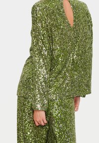 Persona che indossa un abito verde con paillettes, maniche lunghe e pantaloni, mano rilassata al fianco, sfondo chiaro uniforme.