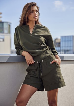 Olivgrünes Oberteil mit langen Ärmeln, V-Ausschnitt und Logo, kombiniert mit passenden Shorts, die einen elastischen Bund und eine Kordel haben.